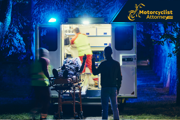 Paramedics load a severely injured patient into an ambulance at night, illuminated by blue emergency lights. The image captures the tragic motorcycle crash on Highway 99 in Merced, where one man died and a woman was critically injured, as reported by Motorcyclist Attorney.